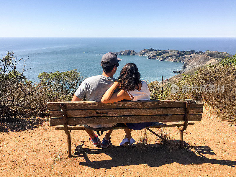 一对年轻夫妇坐在长椅上欣赏美妙的海景