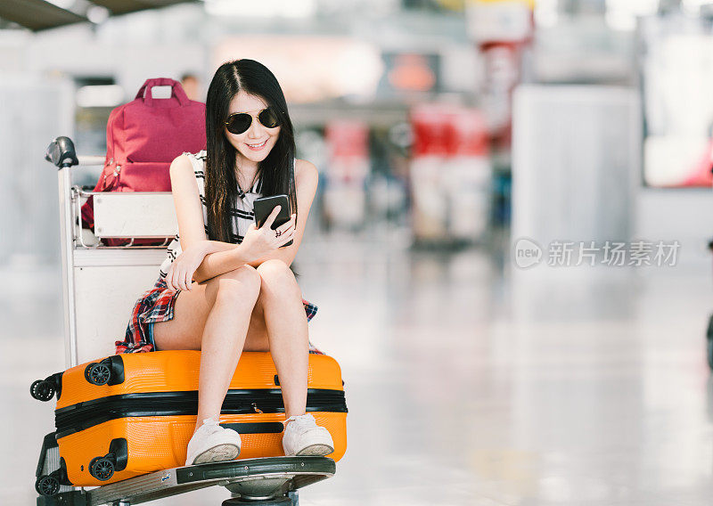年轻的亚洲女孩使用智能手机，在机场等待航班，坐在行李手推车或行李车上。移动通信技术或出国旅游概念，具有复制空间