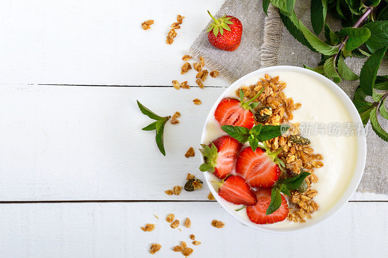 一碗饮食健康早餐:酸奶，格兰诺拉麦片，新鲜草莓，薄荷在白色的木制背景。适当的营养。前视图。