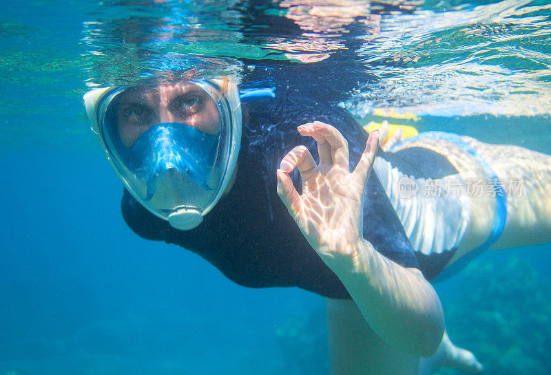 女子浮潜在蓝色的水中，用手指表示ok。戴着面罩浮潜。
