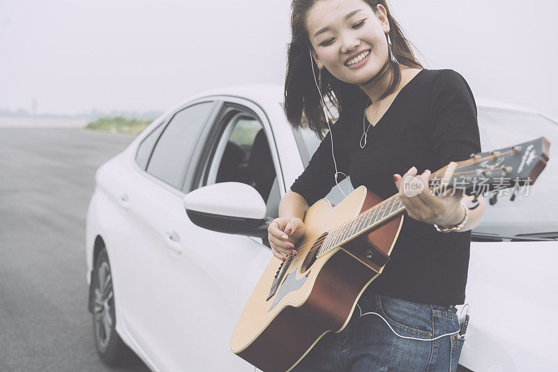 美丽的女孩听着音乐，靠在汽车引擎盖上弹吉他
