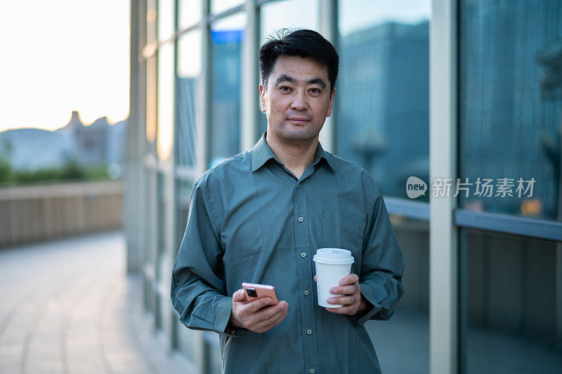 亚洲中年商人拿着咖啡杯，在城市街道上使用智能手机