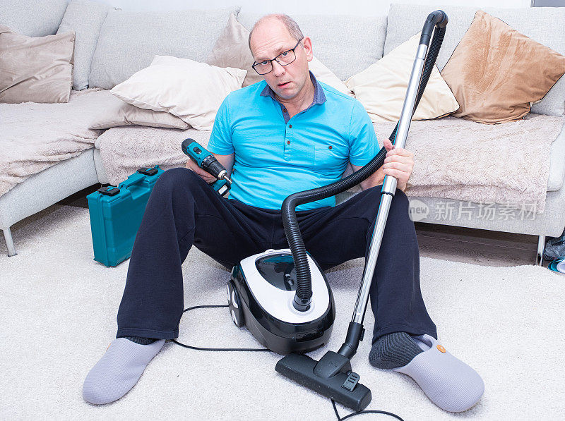 一个白人男人坐在地板上，手里拿着吸尘器和一把无绳螺丝刀。他厌倦了家务活。