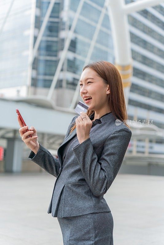 一名年轻的亚洲女商人穿着灰色的办公套装，在商业区用她的智能手机使用信用卡进行在线交易，她微笑着自信