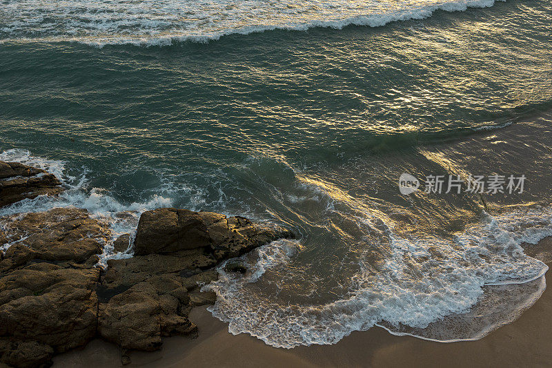 蓝色的海浪拍打着金色的沙滩。太阳照在水面上，石头很大，从上面看。