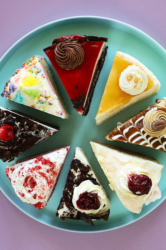粉彩蓝色盘子上各种蛋糕切片的图像，天使食物蛋糕，巧克力和樱桃蛋糕，黑森林蛋糕，红丝绒蛋糕，彩虹蛋糕，咖啡和大理石巧克力蛋糕，盐焦糖蛋糕，紫色背景，高视图