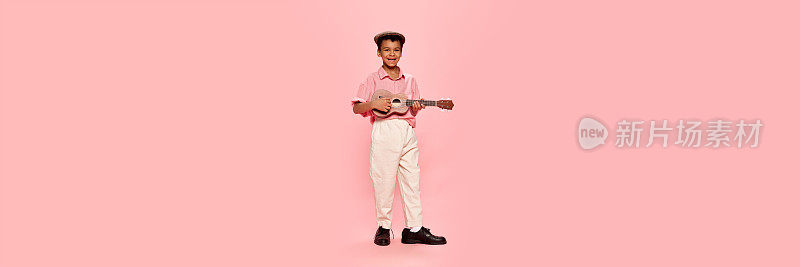 快乐的情感小非洲男孩在复古风格的衣服和帽子摆姿势与尤克里里吉他孤立在粉红色的背景。