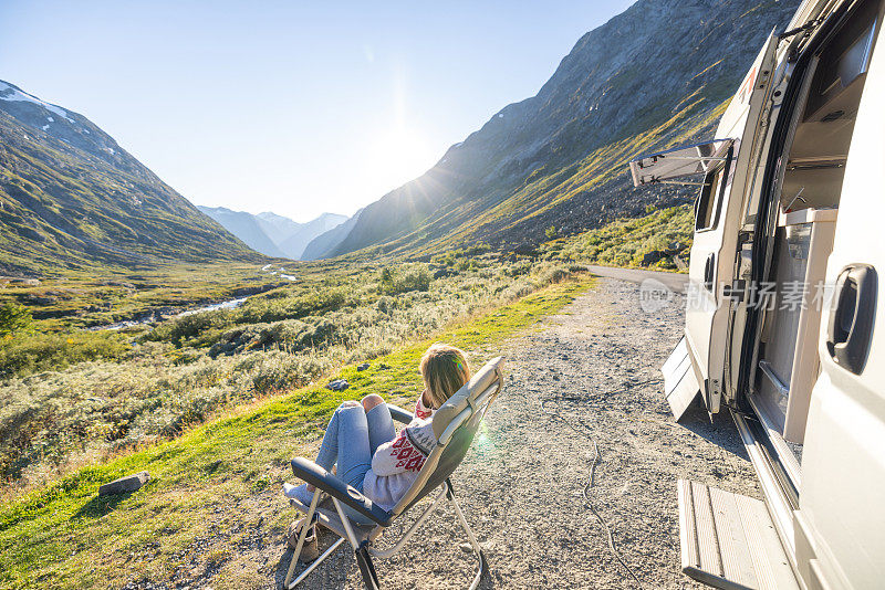 日落时分，女人在她的露营车旁放松，面包车生活的概念。她坐在露营椅上