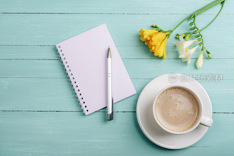 空白记事本，钢笔和咖啡杯放在蓝绿色的木桌上，桌上有小苍兰花。俯视图，平面布局，模型