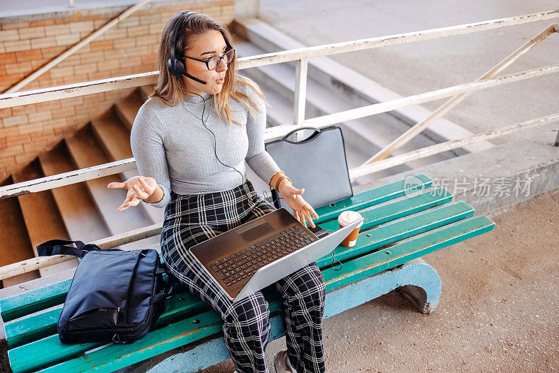 在上班的路上等火车的时候，年轻女子坐在长凳上用笔记本电脑开在线会议