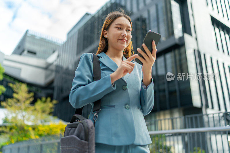 移动中的商业。连接移动。一位年轻的亚洲女商人背着笔记本电脑包，用智能手机上下班，她在中央商务区上班，对面是公司大楼