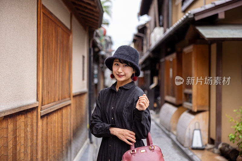 身穿黑色连衣裙、头戴帽子的年轻女子站在日本传统的狭窄街道上的肖像