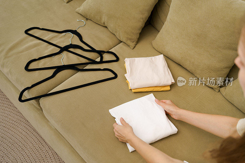 这个女人把沙发上从衣架上取下的t恤整齐地折叠起来。衣橱季节变化的概念，衣柜的清洁和整洁
