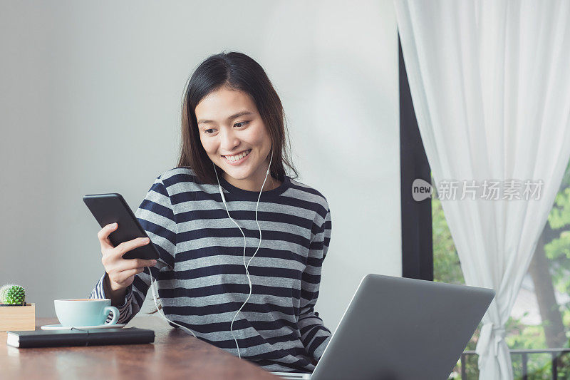 快乐的亚洲女人使用手机聊天，使用笔记本电脑和听音乐，而工作在咖啡馆餐厅的窗口附近，数字时代的生活方式，工作在办公室之外的概念