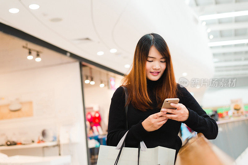 肖像美丽的年轻亚洲女人在购物中心，微笑使用智能手机网络室内。女游客利用科技，旅行生活。购物中心外设有服装店。