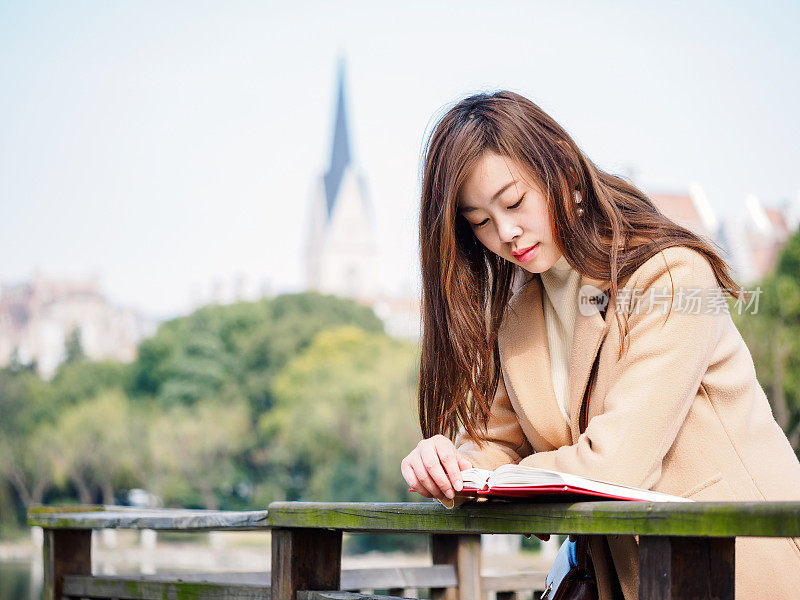 在模糊的哥特式建筑背景下，美丽的中国女孩站在风中读书。大学里的好读者。