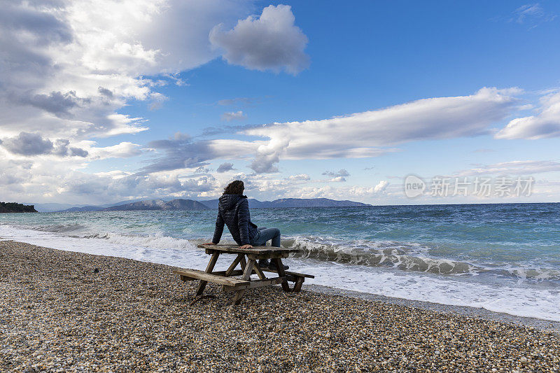 男人看着波涛汹涌的大海，坐在海滩上的野餐桌上，波涛汹涌的大海，大风的天气