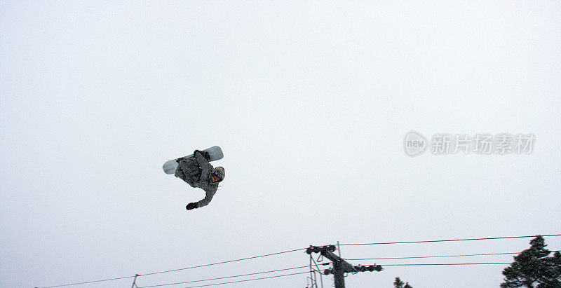 在科罗拉多州博尔德附近的埃尔多拉滑雪度假村，在一个多雪的阴天，一个滑雪板在森林和滑雪缆车的背景下尝试一个“后空翻屈膝”跳跃技巧
