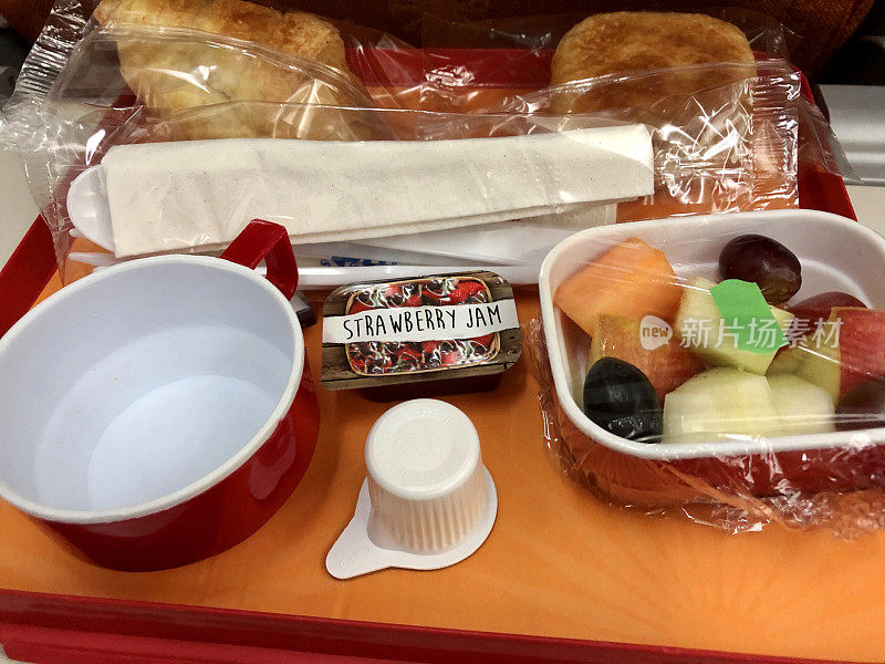 飞机餐盘的特写图片，里面有羊角面包，草莓酱，一碗水果沙拉和一个丹麦苹果，一次性塑料餐具在玻璃纸上，俯瞰