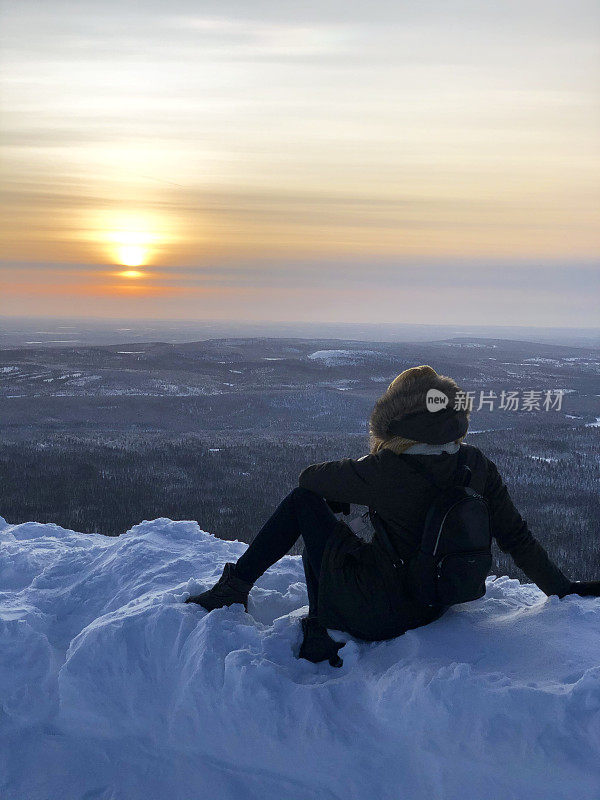 一个年轻的女人坐在山顶上欣赏日落。拉普兰的雪坡。多雪，冬天的风景和很多雪。