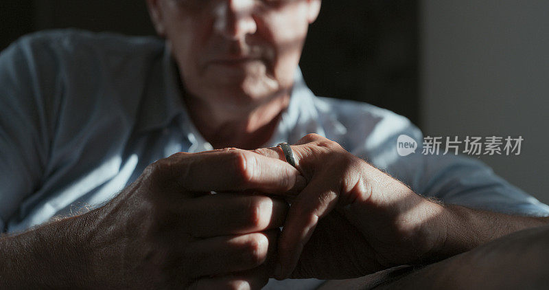 一个不认识的男人独自坐在家里的一个黑暗的房间里，沉思地看着他的结婚戒指