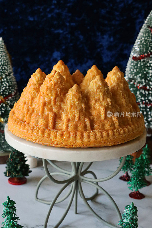 自制的圣诞树形状的柠檬毛绒绒的Bundt蛋糕，放在白色、大理石和金属的蛋糕架上，周围环绕着圣诞树模型，柠檬味的环状海绵覆盖着糖霜，蓝色背景