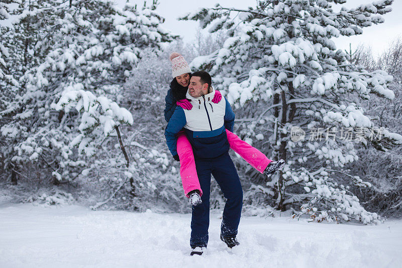 年轻的男朋友背着他的女朋友在雪地里玩，周围是雪树