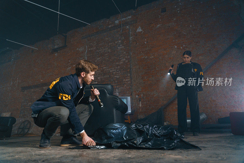 两名FBI探员在犯罪现场工作，其中一名探员正在检查躺在地上黑色塑料袋下的受害者的脉搏
