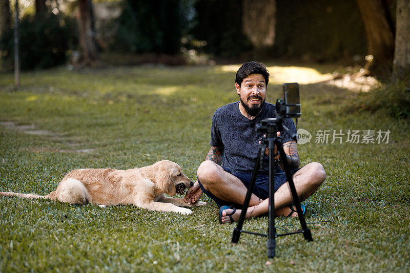 一名男子坐在草地上拍摄自己，一只金毛猎犬小狗在玩松果