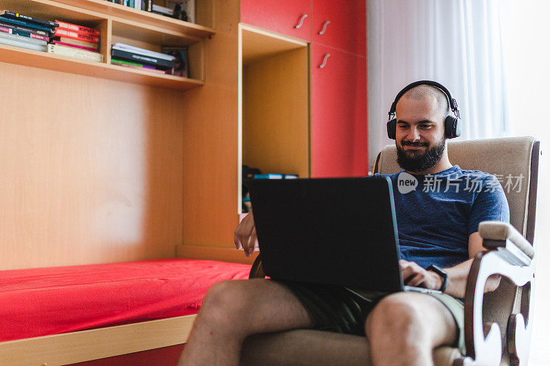 快乐的秃头年轻人在家里戴着耳机用笔记本电脑