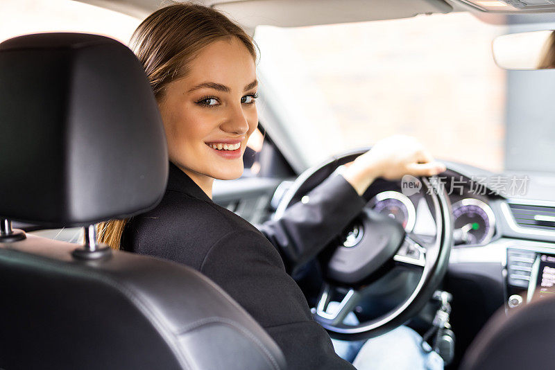 年轻漂亮的黑发女子坐在一辆现代汽车的驾驶座上，握着方向盘转过身，微笑着看着镜头。旅游的概念