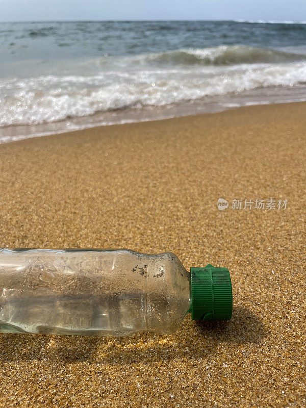 特写图像，空的，透明的塑料饮料瓶与绿色盖子被冲到沙滩上的退潮水的边缘，破碎的海浪冲刷海洋垃圾和岸上的污染，乱扔的沙子，肮脏的海滩，关注前景，复制空间