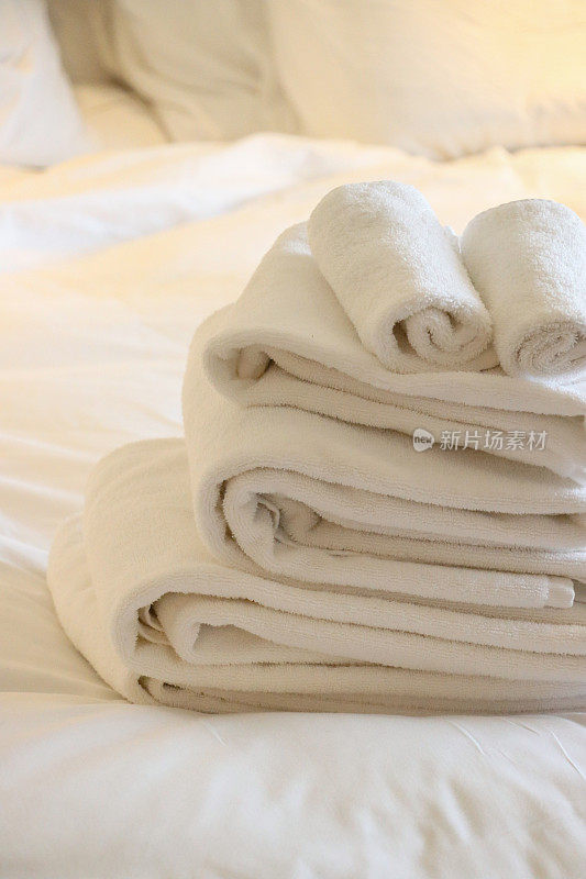 特写图像的堆叠白色酒店毛巾坐在白色羽绒被双人床盖，折叠浴床单和手巾，堆白色枕头背景，重点在前景