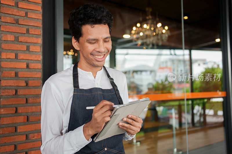 亚洲男子咖啡师手臂使用数码平板电脑接收顾客的订单，并微笑着倚靠在咖啡店门前的玻璃门上