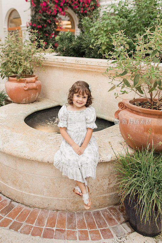 2023年5月，在佛罗里达州棕榈滩的一个托斯卡纳风格的花园中，一个4岁的古巴裔美国女孩，棕色卷发，穿着鼠尾草绿和白色的春季连衣裙，扎着发结，穿着白色的皮凉鞋