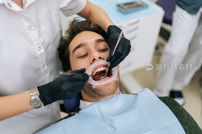 在牙科诊所里，一位不知名的女牙医用牙科工具检查牙齿时，英俊的男性病人张开嘴坐在口腔医生的座位上。
