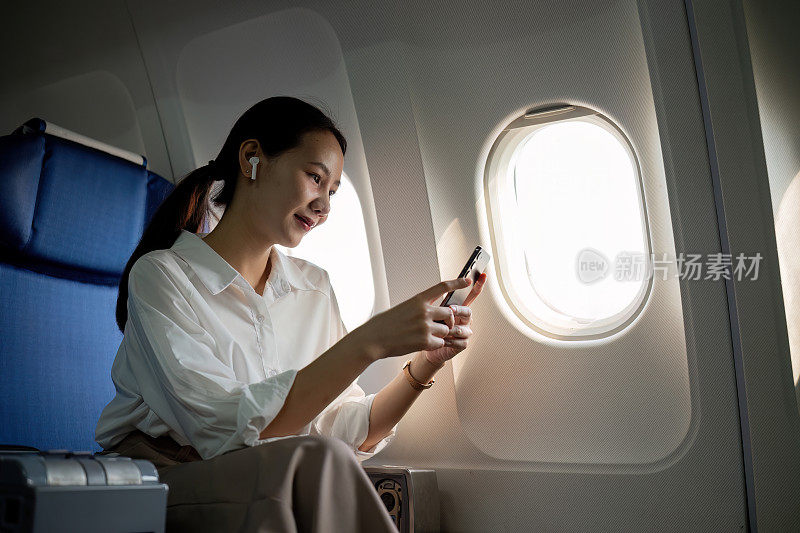 年轻的亚洲女性用手机查看新闻信息，坐在商务舱靠窗的位置，飞行时的飞机座位，旅行和商业概念