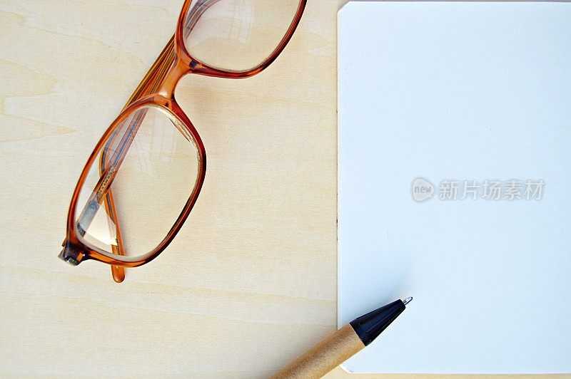 一张水平的眼镜照片，一支钢笔和一张白色的笔记本纸，在一个木制的米色水平背景上很有美感地排列着。