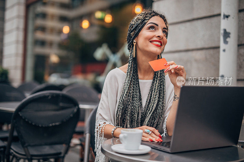 一位女士坐在路边的咖啡馆里，拿着信用卡使用笔记本电脑