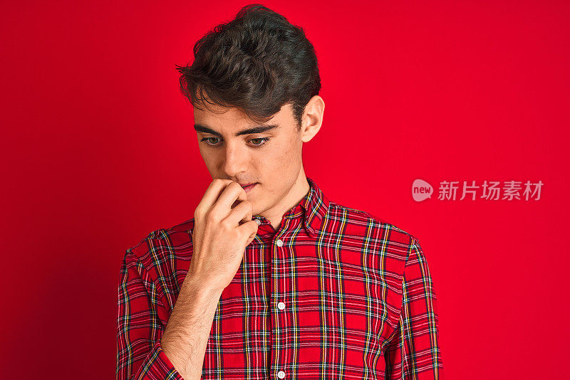 十几岁的男孩穿着红色衬衫站在孤立的背景看起来紧张和紧张的手在嘴咬指甲。焦虑的问题。