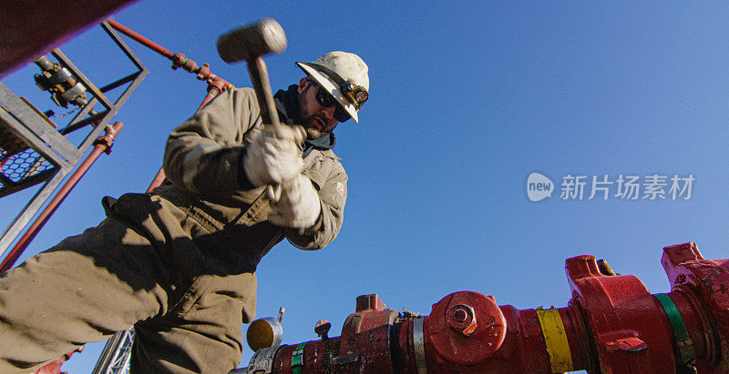 一个寒冷，阳光明媚的冬天早晨，一个30多岁的油田工人在石油和天然气钻井平台上用锤子敲击管道