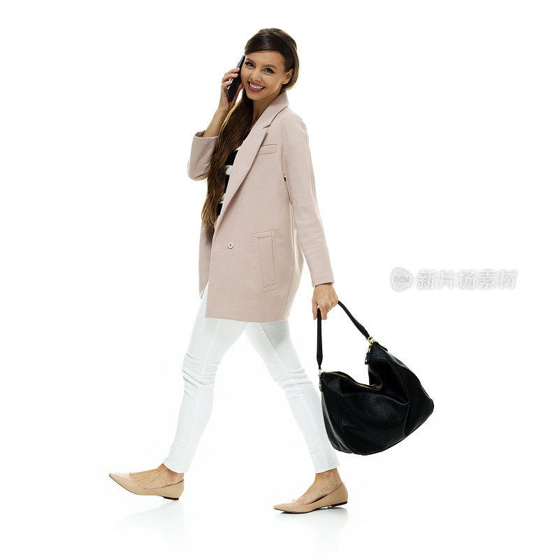 白人女性穿着夹克，拿着钱包，使用智能手机走在白色背景前