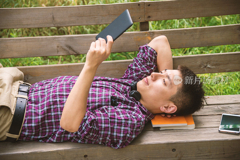 英俊的亚洲男人躺在长凳上阅读数字平板电脑