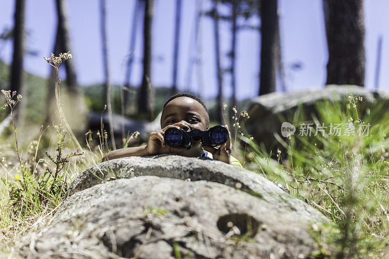 一个非洲男孩拿着望远镜躲在石头后面