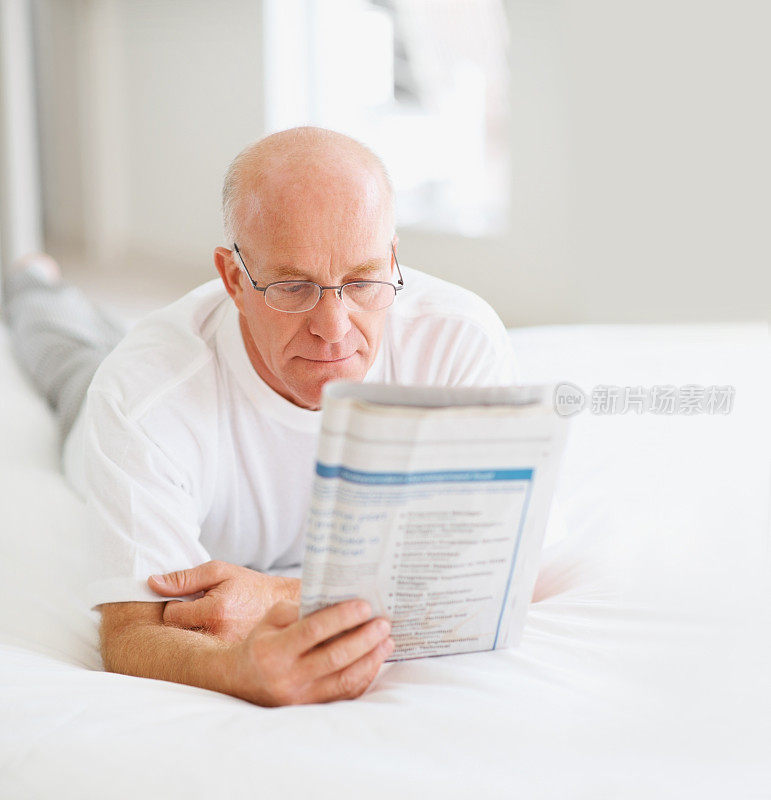 老人躺在家里的床上看报纸