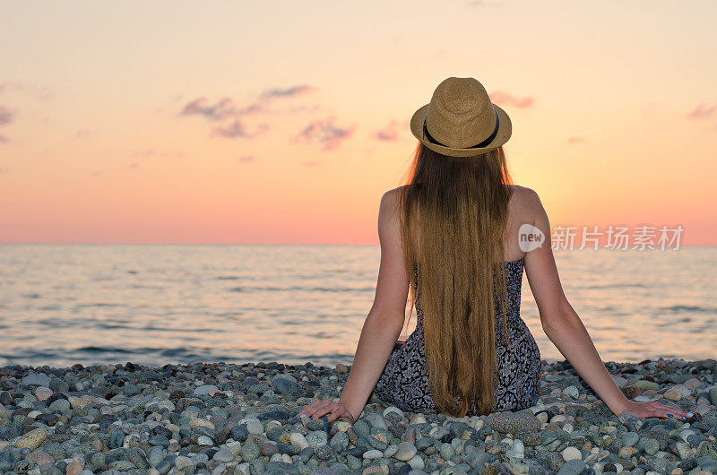 戴着帽子的女孩坐在海边。日落时间。从后面看