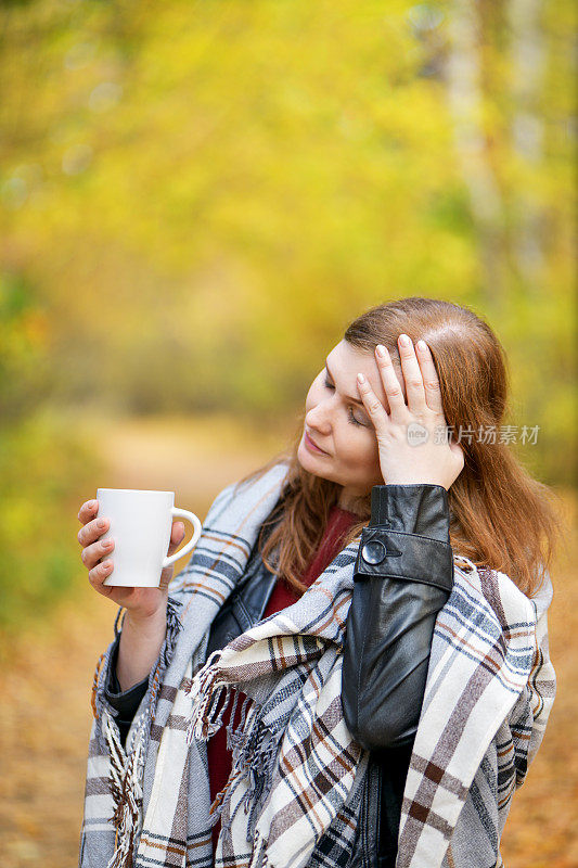 保持你的头。秋天。一个美丽的红发女孩站在秋天的森林里，端着一杯热茶，抱着她的头，裹在一件格子花呢里。秋天的背景。垂直摄影