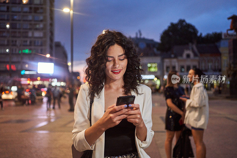 美丽的年轻卷发女人在晚上在户外用手机发短信