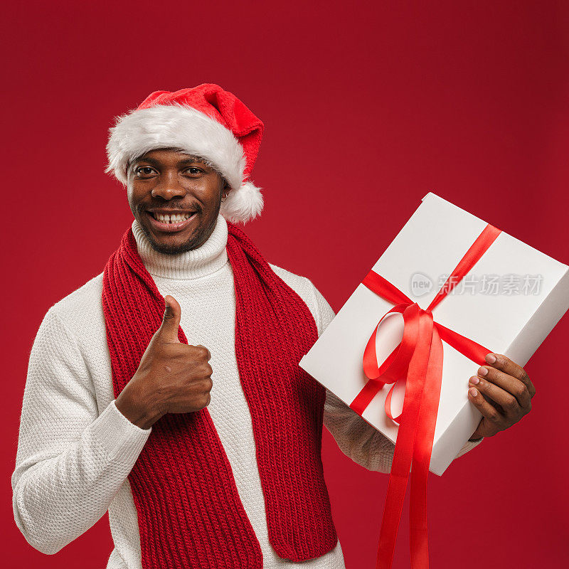 一个穿着毛衣、戴着圣诞帽、皮肤黝黑的男人拿着一个礼盒，竖起大拇指