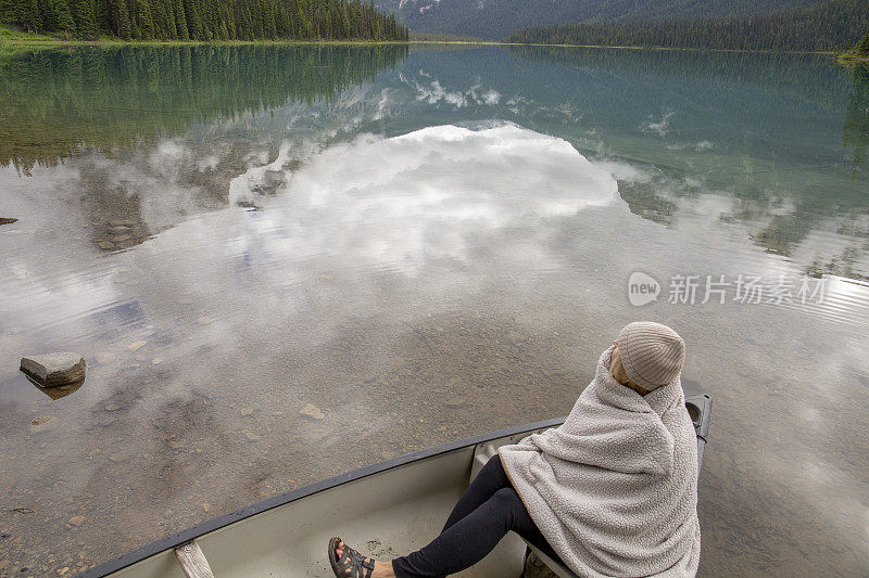 一个裹着毯子的女人坐在高山湖上的小船上